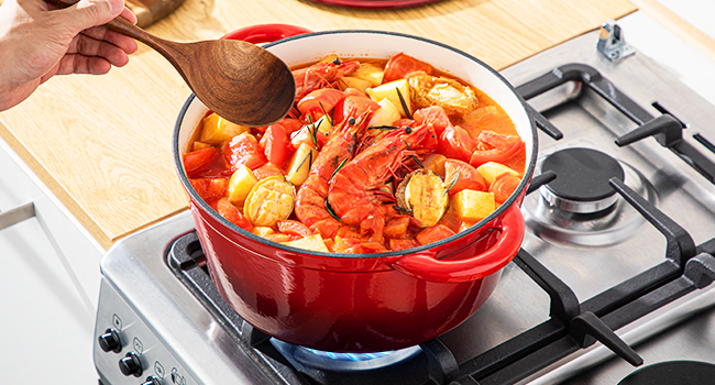 Four hollandais émaillé rouge rempli de fruits de mer bouillis contenant des crevettes, du maïs et des légumes cuits sur une cuisinière à gaz, avec une cuillère en bois en remuant