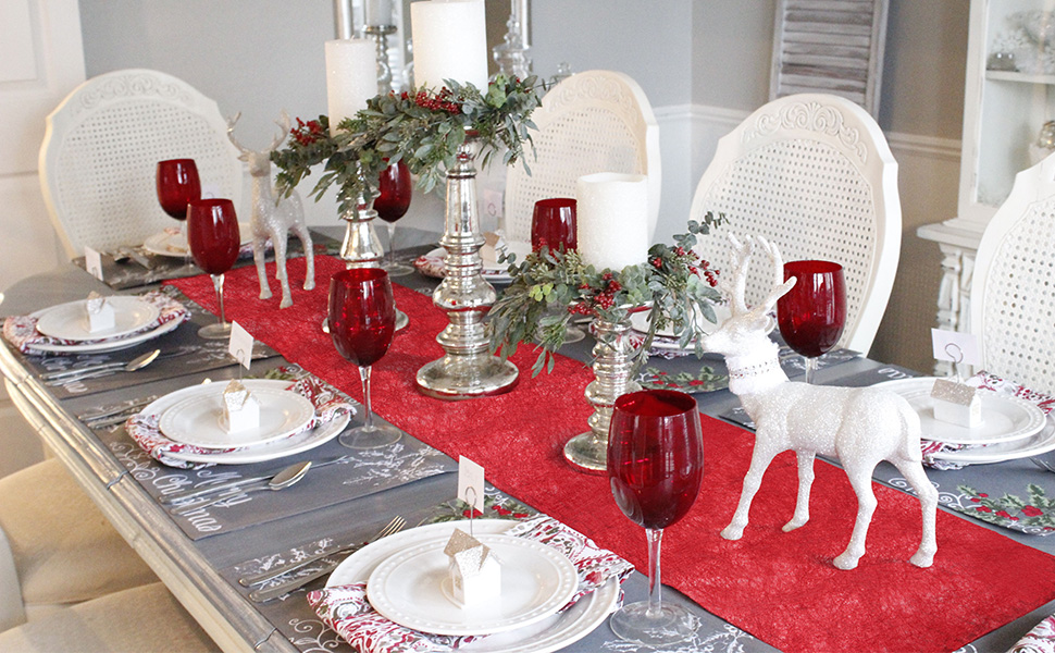 Décoration de table de Noël élégante avec chemin rouge, assiettes blanches, verres à vin rouge, verdure et figurine de renne blanc. Des chaises en osier blanc entourent la table à manger festive
