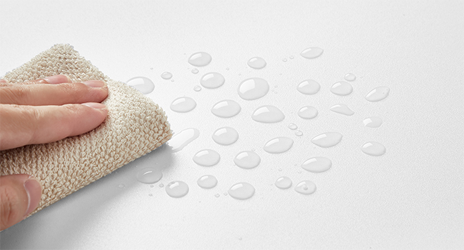 Utilisez à la main un chiffon beige pour essuyer les gouttelettes d'eau sur une surface lisse et blanche, démontrant ainsi des propriétés hydrofuges ou faciles