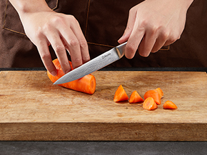 Les mains utilisent un couteau de chef pour trancher une carotte sur une planche à découper en bois, démontrant ainsi une technique de coupe précise.