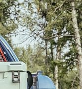 Vue partielle d'une glacière ou d'un box de camping de couleur claire muni d'une poignée, à côté d'une surface bleue, peut-être d'un véhicule. Arbres visibles en arrière-plan.