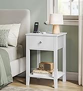 Table de chevet en bois blanc avec tiroir simple et étagère inférieure, placée à côté d'un lit. Lampe de table et appareil électronique visibles sur le dessus.