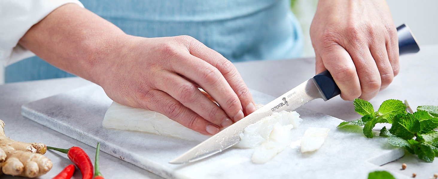 Les mains utilisent un couteau de chef pour hacher des herbes fraîches sur une planche à découper blanche, les légumes étant visibles dans le cadre.
