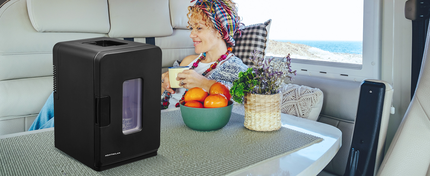 Réfrigérateur noir compact sur une table à l'intérieur d'un véhicule. Bol de fruits et verre à boire à proximité, vue sur la mer par la fenêtre.