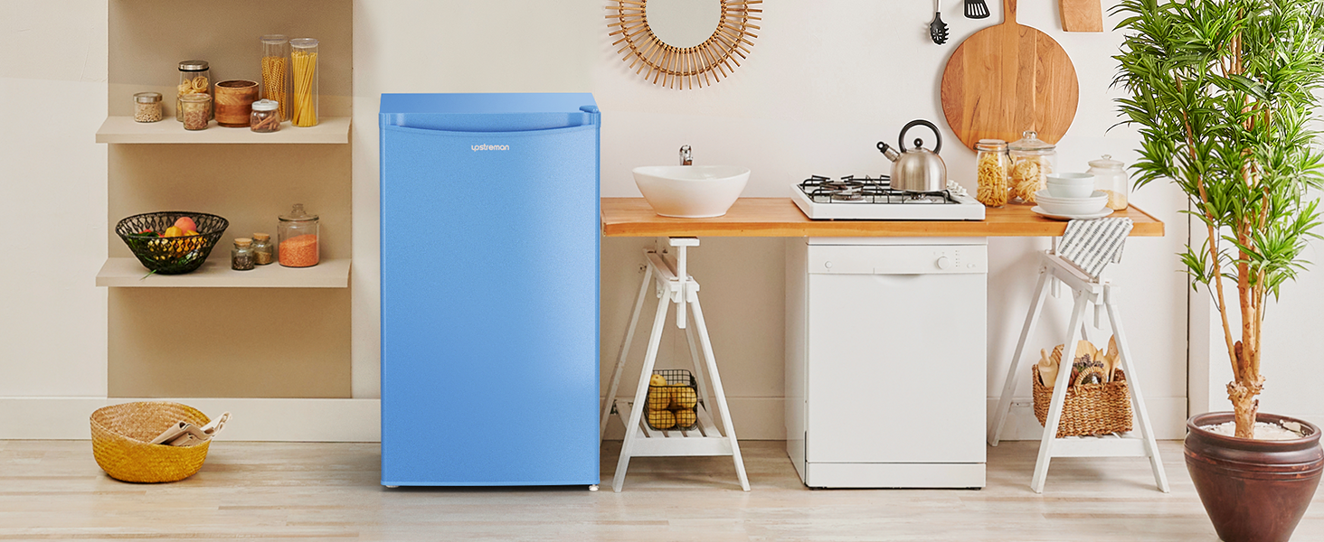 Réfrigérateur bleu compact dans une cuisine. Comptoirs en bois, armoires blanches et étagères avec ustensiles de cuisine visibles. Le petit électroménager et les objets de décoration complètent le décor.