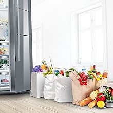 Réfrigérateur en acier inoxydable à côté de plusieurs sacs d'épicerie en papier blanc remplis de produits frais, de fruits, de légumes et de pains.