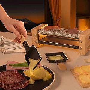 Scène de cuisine montrant des mains utilisant un appareil à raclette en bois pour faire fondre du fromage, avec de la viande et des légumes dans des assiettes sombres à proximité. Configuration traditionnelle de la raclette sur une surface en bois.