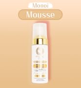 Flacon cylindrique blanc aux accents dorés contenant de la mousse au Monoï. Le produit est doté d'un distributeur à pompe et comporte des éléments décoratifs dorés sur l'étiquette.