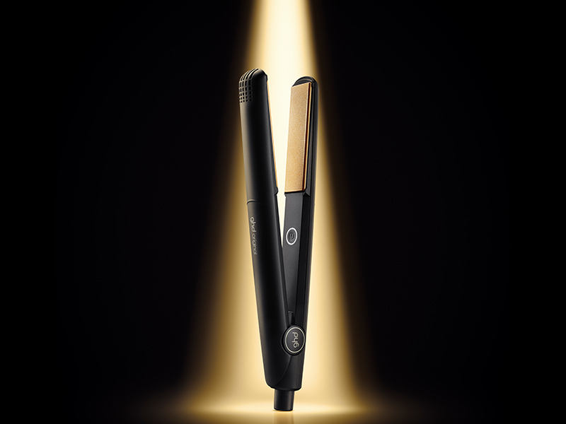 Un fer à lisser ou un fer à coiffer noir s'éclaire de façon spectaculaire sur fond sombre avec un effet de lumière dorée derrière lui.