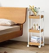 Chariot roulant à trois niveaux avec étagères en bois et structure en métal blanc, placé à côté d'un lit. L'étagère supérieure contient des fleurs, l'étagère du milieu contient des bouteilles de cosmétiques, l'étagère inférieure contient des serviettes pliées.