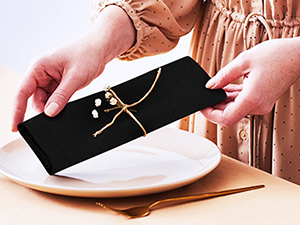 Des mains tenant une boîte rectangulaire noire avec des détails dorés sur une plaque blanche, suggérant un emballage de bijoux ou d'accessoires.