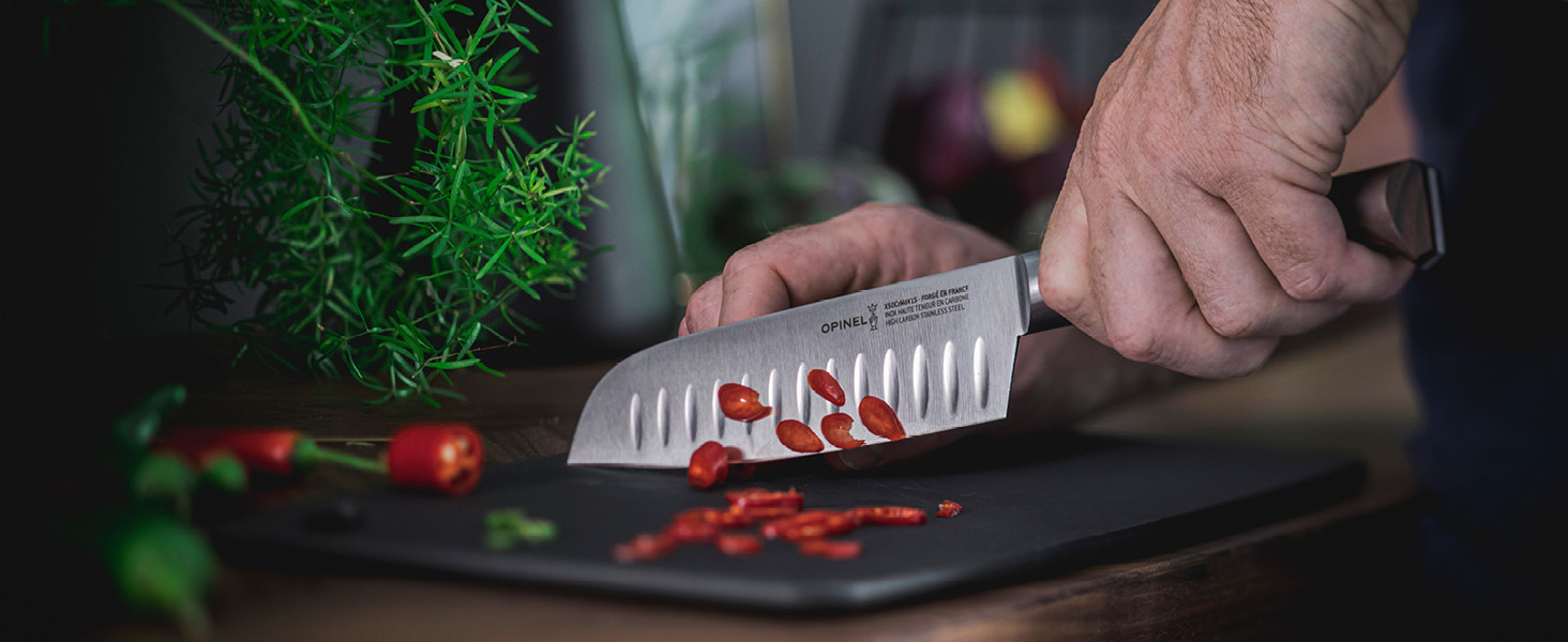 À la main, tenant un couteau de chef, tranchant des piments rouges sur une planche à découper foncée. Herbes fraîches visibles en arrière-plan.