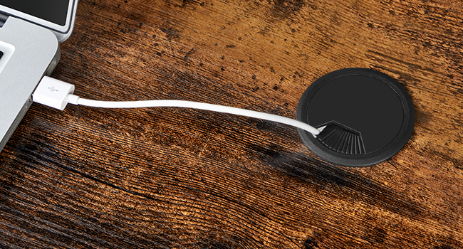 Appareil circulaire noir avec structure en forme de ventilateur connecté à un câble blanc, posé sur une surface en bois à côté du port USB de l'ordinateur portable