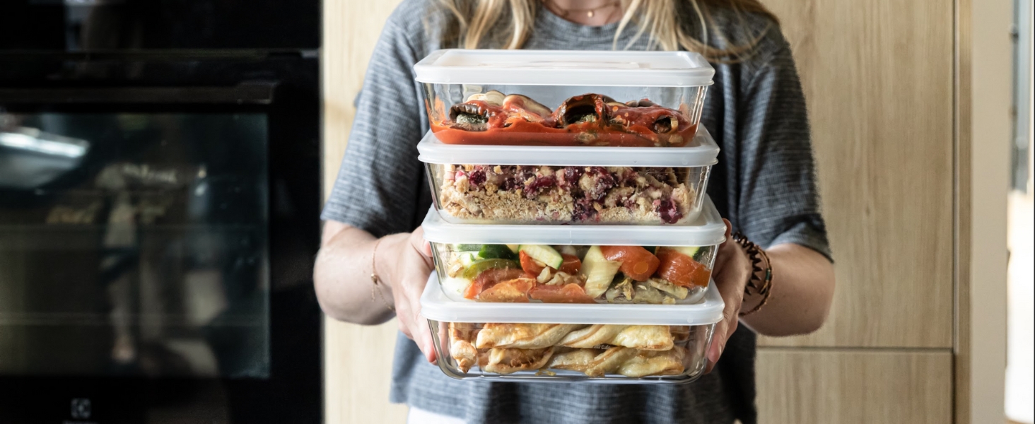 Les mains tiennent une pile de contenants alimentaires en plastique transparent contenant divers plats préparés à l'intérieur, pour démontrer la préparation des repas ou la conservation des aliments.