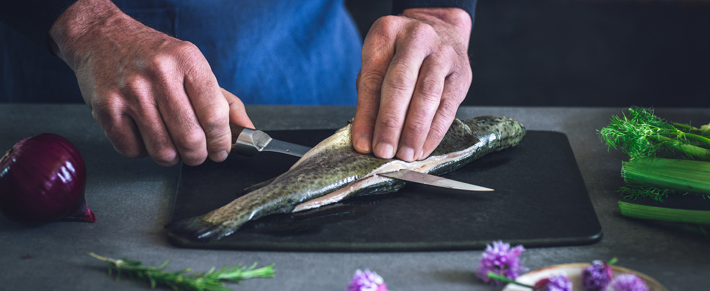 Les mains utilisent un couteau pour préparer le poisson sur une planche à découper noire. Les autres ingrédients visibles incluent l'oignon rouge et les herbes fraîches.