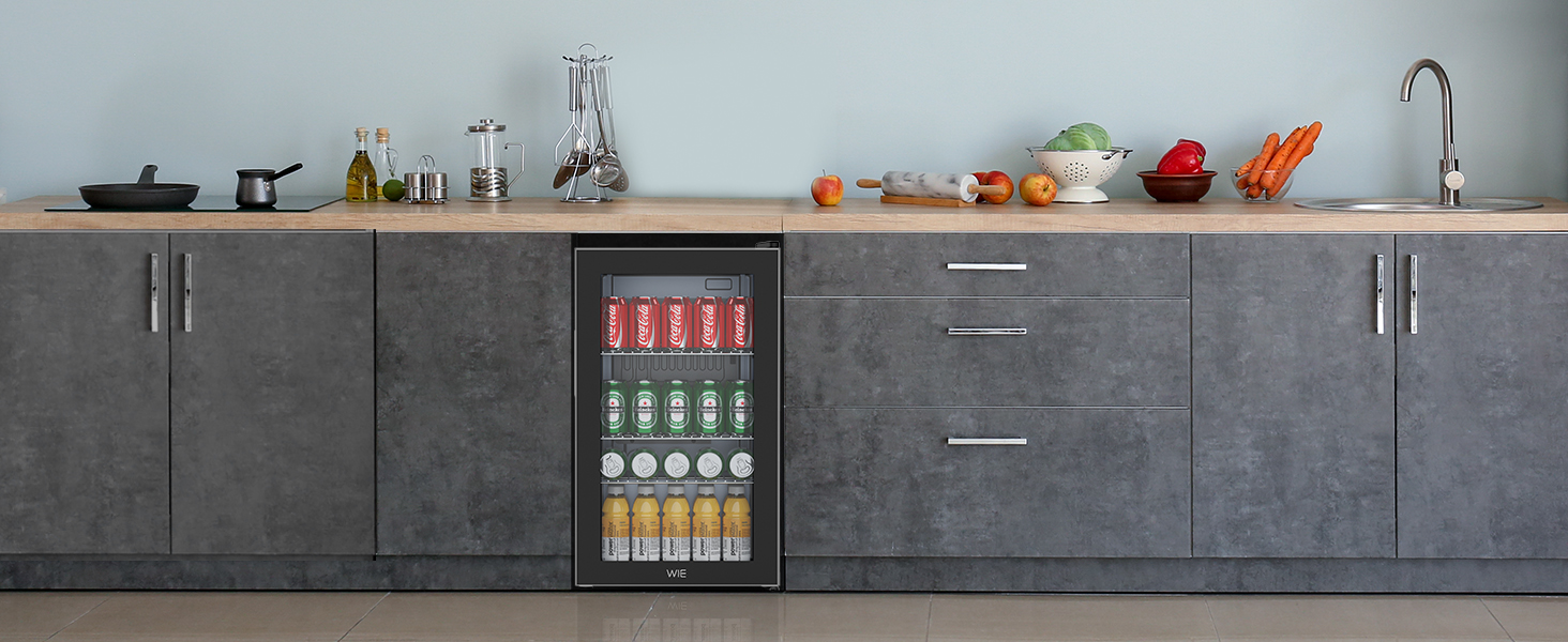 Cuisine moderne avec armoires grises, comptoir en bois et appareils électroménagers en acier inoxydable. Petit réfrigérateur à boissons visible. Divers produits alimentaires et ustensiles sur le comptoir.