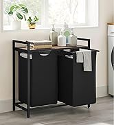 Chariot à linge en métal noir avec deux compartiments en tissu et une étagère supérieure en bois. Placé près d'une fenêtre avec des plantes et des accessoires de salle de bain visibles.