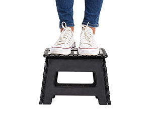 Pieds en baskets blanches debout sur un petit escabeau pliable noir sur fond blanc.