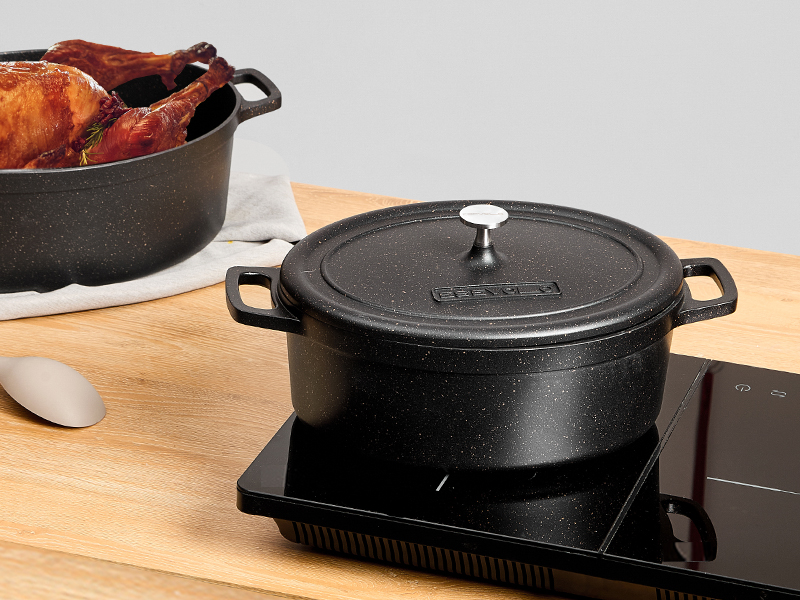 Four hollandais en fonte noire avec couvercle posé sur une planche à découper en bois, illustré à côté d'un autre récipient de cuisson contenant des aliments. Four hollandais placé sur ce qui semble être une table de cuisson à induction.