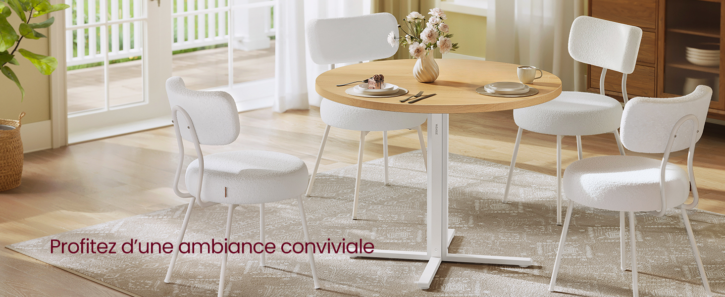 Table à manger ronde en bois avec pieds blancs, entourée de quatre chaises blanches. Set de table avec assiettes et fleurs, dans une pièce lumineuse avec fenêtres.