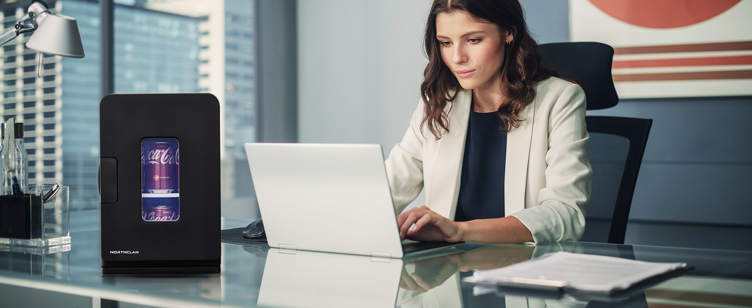 Femme utilisant un ordinateur portable au bureau avec un petit appareil noir affichant une lumière violette. Cadre de bureau avec stores visibles en arrière-plan.